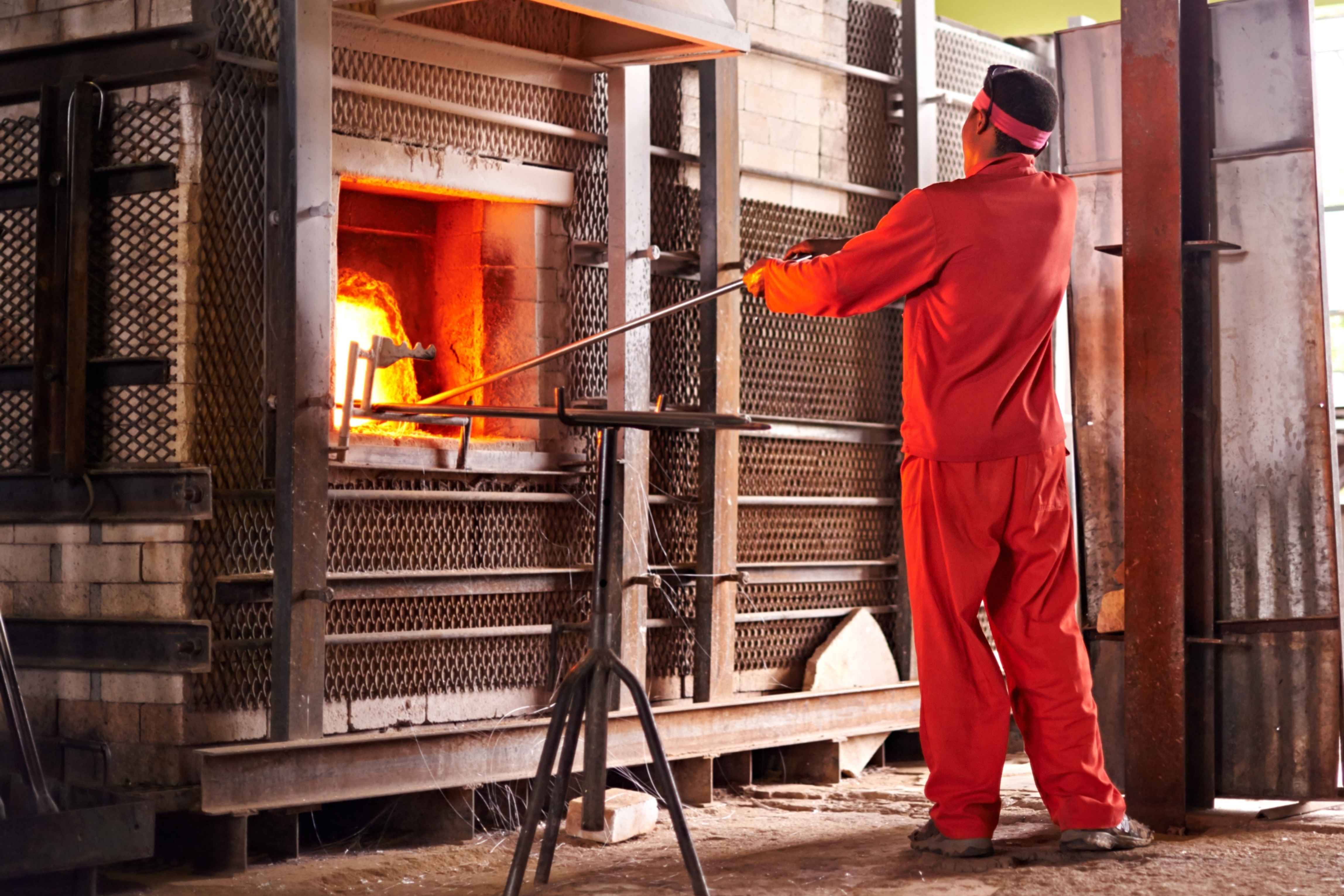 Glass factory, man and fire in furnace for production process, manufacturing and back in industry. Glassware, tools and craft for art with worker for recycling, melt and African person glassblowing